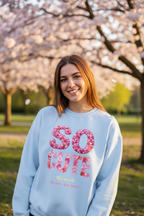 Woman wearing a light blue sweatshirt with 'So Cute' print, standing in a park with cherry blossom trees.