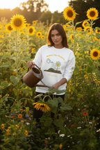 Woman holding a watering can in a sunflower field wearing a white sweatshirt with graphics.