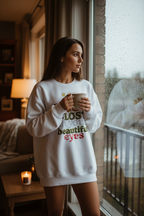 Woman in a white sweatshirt with text holding a mug by a window.
