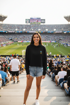 Woman standing on a stadium steps wearing a black sweatshirt and denim shorts, with a sports event in the background.